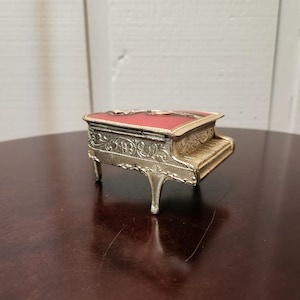 Könnte beinhalten: Eine Miniatur-Spieldose in Form eines Klaviers mit roter Oberseite und goldfarbenen Akzenten. Das dekorative Klavier weist verzierte Details an den Beinen und Seiten auf. Die Tasten sind hellgoldfarben. Die Spieluhr ist ein dekorativer Gegenstand.