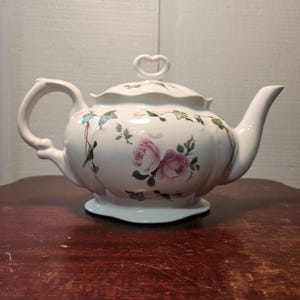 Puede incluir: Una tetera de cerámica de color crema con un delicado patrón floral de rosas rosadas y enredaderas de hiedra verde. La tetera tiene una base festoneada, un asa curva, un pico cónico y una tapa rematada con un asa en forma de corazón.