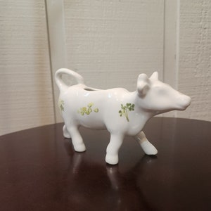May include: White ceramic creamer in the shape of a cow with green and yellow floral designs.