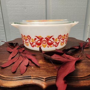 May include: White casserole dish with a clear glass lid. The dish features a decorative pattern of red and orange birds and floral designs. The dish is sitting on a wooden surface.
