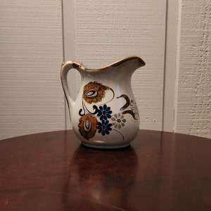 May include: A white ceramic jug with a brown handle and a floral design in shades of blue, brown, and grey.