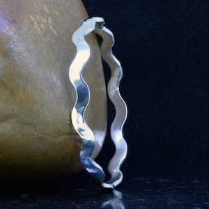 May include: A silver-coloured, wavy bangle bracelet. The bracelet has a unique, undulating design, with a smooth, polished finish. The bracelet is displayed against a dark background, highlighting its reflective surface. The bracelet is a piece of jewellery.