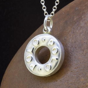 May include: A silver-colored pendant necklace with a circular charm. The charm has the word "POLO" embossed around the edge. The pendant hangs from a delicate silver chain. The necklace is displayed on a brown surface.