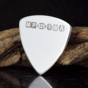 May include: A shiny, silver-coloured guitar plectrum with a smooth surface and rounded edges. The pick has the markings "MP 925" and other symbols. It is set against a dark background with a piece of wood.