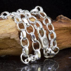 May include: A silver chain bracelet with large, rounded links and a secure clasp. The bracelet is made of shiny metal and is displayed on a piece of wood. The clasp features a decorative emblem.