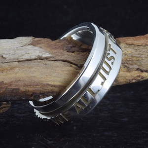 May include: A silver ring with the engraved words "WE ALL JUST" in a bold font. The ring has a polished finish and a simple, elegant design. The ring is displayed on a piece of wood.