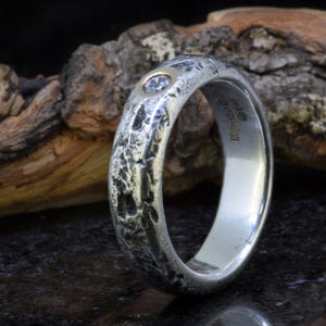 May include: A silver ring with a heavily textured, rugged exterior surface and a single round, clear gemstone set in a gold-toned bezel. The interior of the band features visible hallmarks and stamps, indicating its metallic composition and origin.