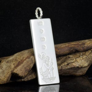 May include: A rectangular silver pendant with a twisted loop at the top. The pendant features an engraved image of a saint carrying a child through water. Hallmarks are visible on the front.
