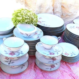 Puede incluir: Un conjunto de vajilla de porcelana vintage con un diseño de rosas. El juego incluye platos, platillos, tazas de té, cuencos y una salsera. La vajilla es blanca con un borde azul claro y un patrón de rosas rosas.