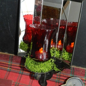 Puede incluir: Un espejo de pared de metal negro con un diseño decorativo. El espejo tiene cuatro paneles de vidrio y está decorado con candelabros de vidrio rojo. Los candelabros están llenos de cera roja y están sentados en un lecho de musgo verde.