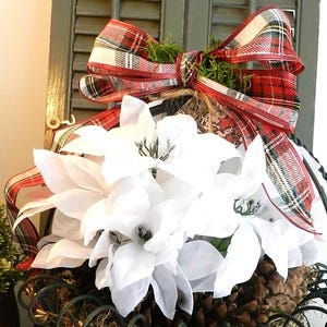 Puede incluir: Una decoración navideña festiva con un gran lazo a cuadros rojo, blanco y verde sobre flores de pascua blancas y piñas. El arreglo se acentúa con vegetación y una estética rústica y natural.