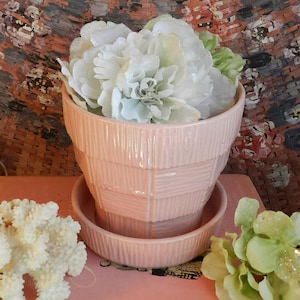 Könnte beinhalten: Ein rosa Keramik-Blumentopf mit passender Untertasse, gefüllt mit weißen und grünen Kunstblumen. Der Topf hat ein strukturiertes, geometrisches Design. Die Anordnung steht auf einer rosa Oberfläche, mit einem korallenartigen Objekt und zusätzlichen Blumen.