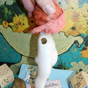 K&ouml;nnte beinhalten: Ein Vintage-Puppenkopf und -torso. Der Puppenkopf ist rot-orange und mit einem rosa Stoff bedeckt. Der Torso ist wei&szlig; mit einem Loch in der Brust. Die Puppe wird von einer Hand gehalten.