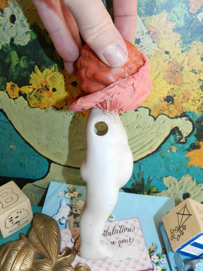 K&ouml;nnte beinhalten: Ein Vintage-Puppenkopf und -torso. Der Puppenkopf ist rot-orange und mit einem rosa Stoff bedeckt. Der Torso ist wei&szlig; mit einem Loch in der Brust. Die Puppe wird von einer Hand gehalten.