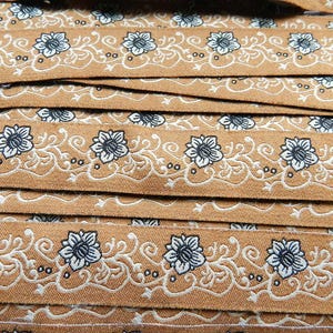 May include: Close-up of brown fabric trim with a white floral pattern. The trim features a repeating design of stylized flowers and vines.