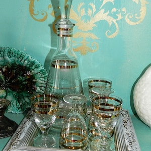 May include: A set of clear glass liquor glasses with gold trim, including a decanter with a gold stopper, on a white framed mirror tray.