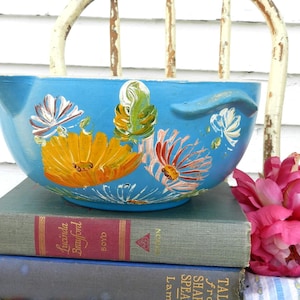 Puede incluir: Un cuenco mezclador de cerámica azul con pico y asa, pintado a mano con diseños florales coloridos. El cuenco descansa sobre una pila de libros antiguos, con una flor rosa en el fondo. Un artículo de cocina encantador y decorativo.