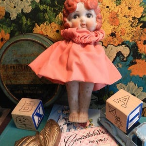 K&ouml;nnte beinhalten: Eine Vintage-Puppe mit roten Haaren und einem rosa Kleid steht vor einem bunten Blumengrund. Holzbuchstabenbl&ouml;cke und eine Gru&szlig;karte befinden sich im Vordergrund. Die Puppe hat ein Porzellangesicht und tr&auml;gt ein rosa Kleid.