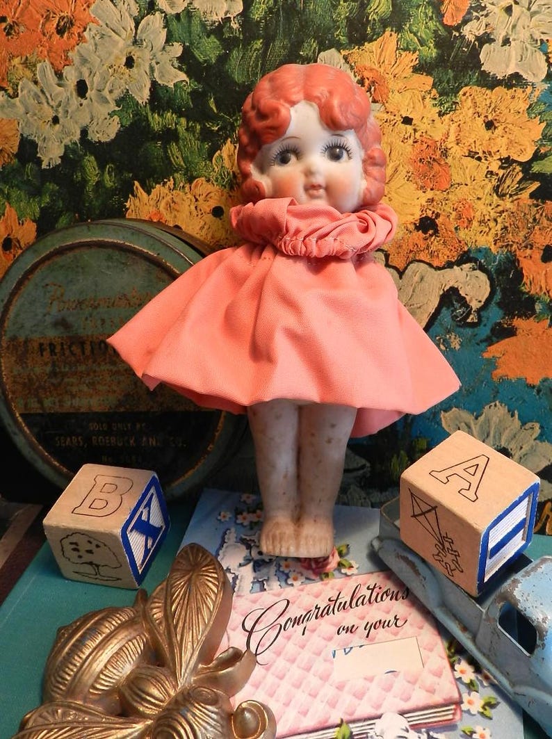 K&ouml;nnte beinhalten: Eine Vintage-Puppe mit roten Haaren und einem rosa Kleid steht vor einem bunten Blumengrund. Holzbuchstabenbl&ouml;cke und eine Gru&szlig;karte befinden sich im Vordergrund. Die Puppe hat ein Porzellangesicht und tr&auml;gt ein rosa Kleid.