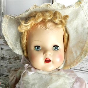 May include: A vintage doll with blonde curly hair, blue eyes, and red lips. The doll wears a pink dress and a white bonnet with lace trim. The doll's face has rosy cheeks and a serene expression, evoking a sense of nostalgia.