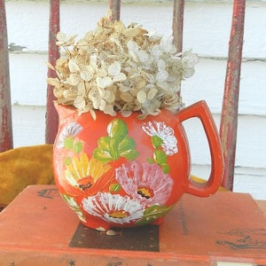 May include: A vibrant orange ceramic pitcher adorned with hand-painted floral designs in white, pink, yellow, and green. The pitcher holds dried white flowers and sits atop a stack of vintage books. The pitcher has a handle.