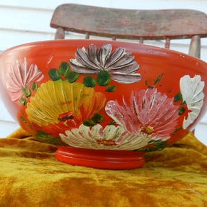 Puede incluir: Un cuenco grande y redondo de madera pintado a mano. El cuenco es de color naranja vibrante con diseños florales coloridos en rosa, amarillo, blanco y verde. El cuenco está sobre una superficie de terciopelo dorado.