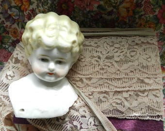 May include: A porcelain doll head with blonde hair and a delicate facial expression. The doll is wearing a lace collar and is displayed on a lace fabric background.