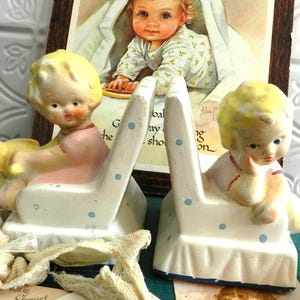 Puede incluir: Un par de sujetalibros de cerámica vintage con figuras de bebés sentados con cabello amarillo y ropa rosa. Los sujetalibros son blancos con lunares azules y una base azul. Un retrato de bebé enmarcado está en el fondo.