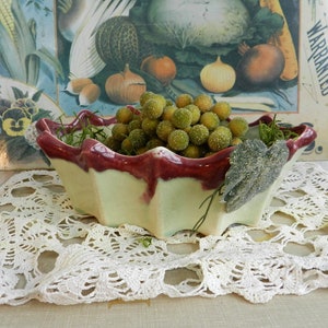 May include: A cream and burgundy ceramic bowl with a scalloped edge, filled with green spheres. The bowl sits on a white lace doily. A vintage vegetable print is visible in the background, creating a rustic aesthetic.