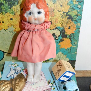 K&ouml;nnte beinhalten: Eine Vintage-Puppe mit Porzellangesicht, rotem Haar und einem rosa Kleid. Die Puppe steht auf einer blauen Karte mit floralem Design. Ein Holzblock mit einer Drachenillustration und ein blaues Spielzeugauto sind ebenfalls zu sehen.