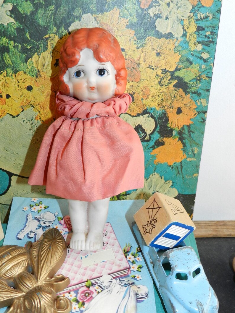 K&ouml;nnte beinhalten: Eine Vintage-Puppe mit Porzellangesicht, rotem Haar und einem rosa Kleid. Die Puppe steht auf einer blauen Karte mit floralem Design. Ein Holzblock mit einer Drachenillustration und ein blaues Spielzeugauto sind ebenfalls zu sehen.