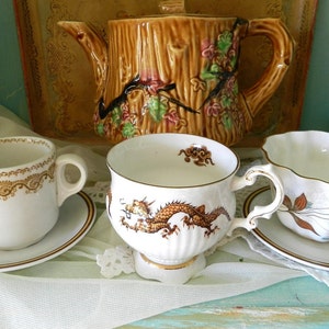 May include: Three teacups and saucers with different designs. One cup features a gold rim and a brown dragon design. Another cup has a gold rim and a brown and gold floral design. The third cup has a gold rim and a brown and white floral design. All three cups are on a white lace tablecloth.