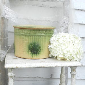 Puede incluir: Un tarro de cerámica vintage, redondo, verde y beige, con tapa, decorado con motivos florales y líneas verticales. El tarro está sobre una mesa blanca desgastada junto a un arreglo de hortensias blancas.