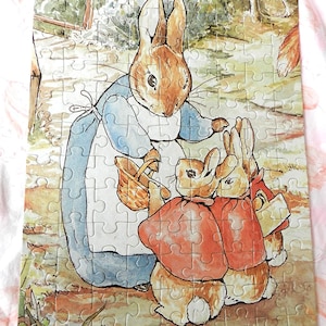 Könnte beinhalten: Eine Aquarellillustration einer Mutterkaninchen, die ein blaues Kleid trägt und einen Korb hält, mit zwei kleinen Kaninchen, die rote Mäntel tragen und vor ihr stehen.