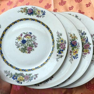 Peut inclure: Ensemble de quatre assiettes en céramique blanche ornées de motifs floraux. Chaque assiette présente un bouquet floral central et une bordure bleue et jaune. Les assiettes ont un bord doré et sont empilées.