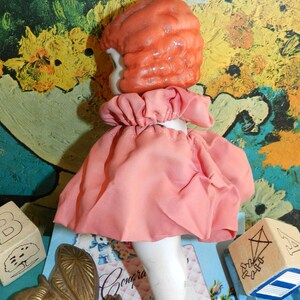 K&ouml;nnte beinhalten: Eine Vintage-Puppe mit pfirsichfarbenem, geformtem Haar und einem rosa Kleid. Die Puppe hat wei&szlig;e Porzellanbeine und einen ger&uuml;schten Kragen. Holzbl&ouml;cke und eine Messing-Schmetterlingsdekoration sind ebenfalls zu sehen.