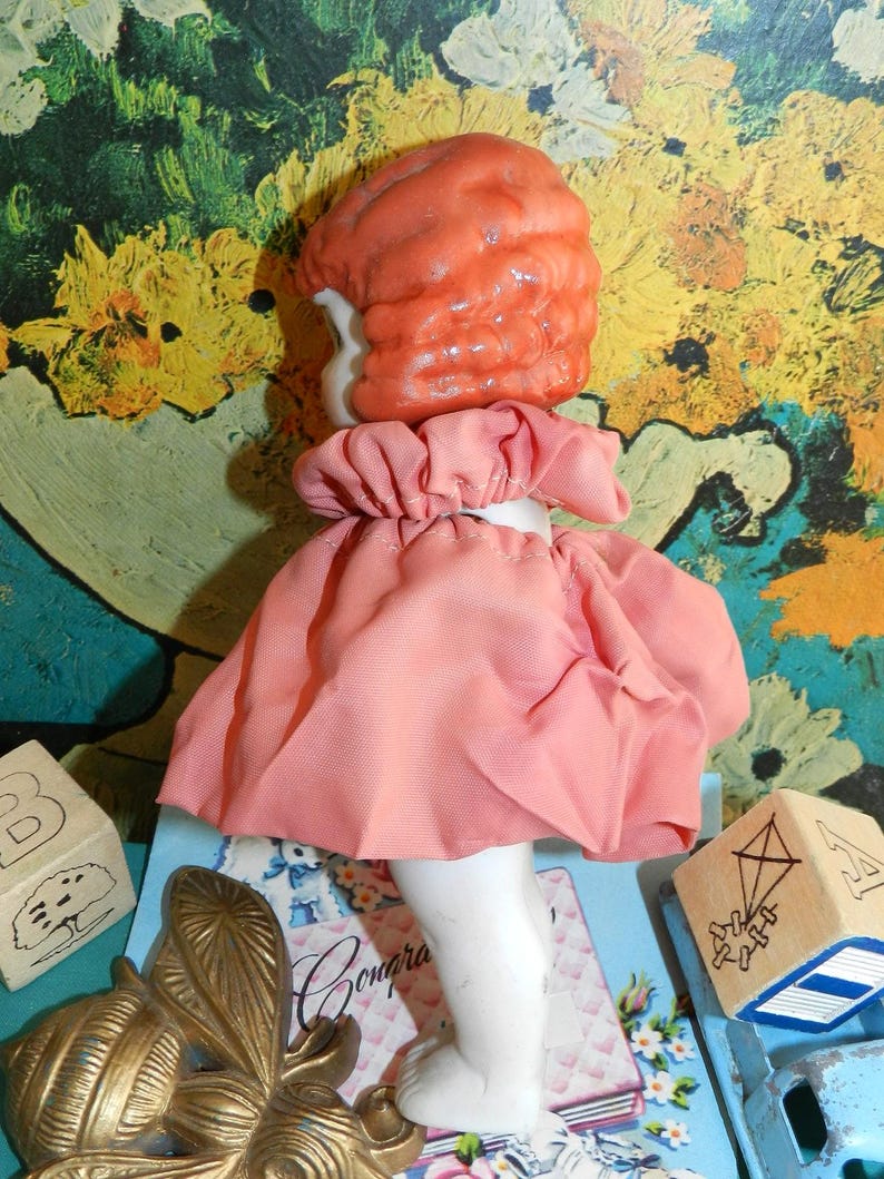 K&ouml;nnte beinhalten: Eine Vintage-Puppe mit pfirsichfarbenem, geformtem Haar und einem rosa Kleid. Die Puppe hat wei&szlig;e Porzellanbeine und einen ger&uuml;schten Kragen. Holzbl&ouml;cke und eine Messing-Schmetterlingsdekoration sind ebenfalls zu sehen.