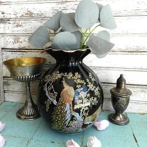 May include: A black ceramic vase with a peacock design, gold trim, and a ruffled top. The vase holds gray faux eucalyptus. A silver goblet and a small silver container are also present, along with pink and white decorative flowers.