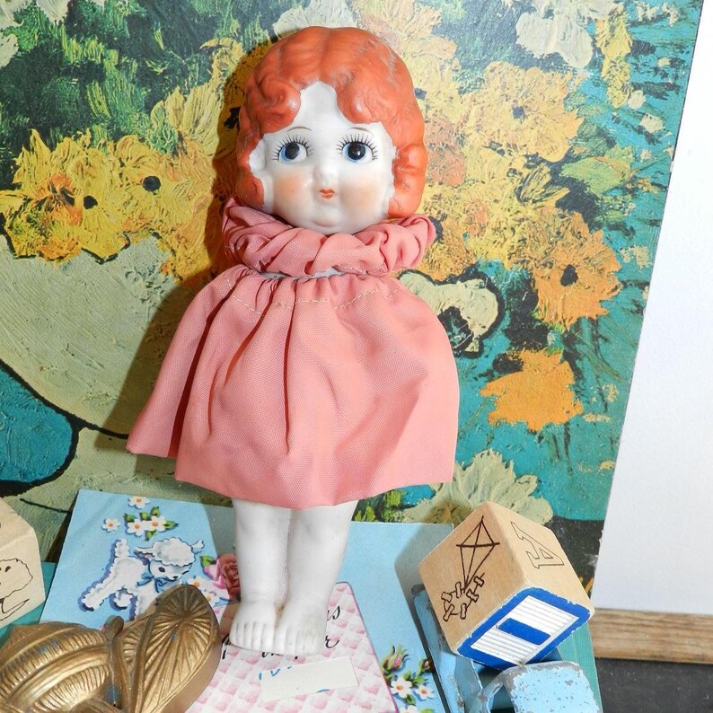 K&ouml;nnte beinhalten: Eine Vintage-Porzellanpuppe mit rotem Haar, blauen Augen und einem rosa Kleid. Die Puppe steht auf einem dekorativen Sockel mit floralem Design. Ein Holzblock mit einer Drachenillustration ist ebenfalls sichtbar.