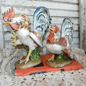May include: Two ceramic rooster figurines, one white with blue and green tail feathers, the other white with brown and blue tail feathers. Both roosters are standing on green bases.