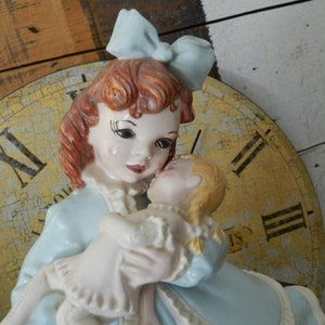 Może przedstawiać: Antyczna figurka porcelanowa przedstawiająca dziewczynkę w niebieskiej sukience trzymającą mniejszą lalkę. Dziewczynka ma rude włosy i niebieską kokardkę. Lalka ma blond włosy i jest ubrana w białą sukienkę.