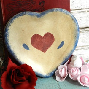 May include: Heart-shaped ceramic plate with a cream-colored center, a red heart, and blue accents. The plate has a blue rim and is surrounded by red roses and pink flowers. A vintage decorative piece.