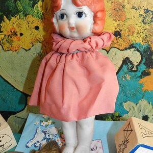 K&ouml;nnte beinhalten: Eine Vintage-Porzellanpuppe mit rotem Haar, blauen Augen und einem rosa Kleid. Die Puppe hat ein rundes Gesicht und steht. Der Hintergrund zeigt ein farbenfrohes abstraktes Gem&auml;lde und anderes Vintage-Spielzeug.