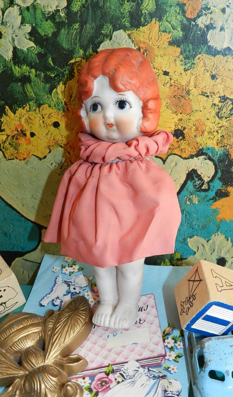 K&ouml;nnte beinhalten: Eine Vintage-Porzellanpuppe mit rotem Haar, blauen Augen und einem rosa Kleid. Die Puppe hat ein rundes Gesicht und steht. Der Hintergrund zeigt ein farbenfrohes abstraktes Gem&auml;lde und anderes Vintage-Spielzeug.