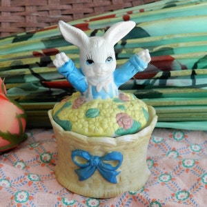 May include: A ceramic trinket box with a white bunny figurine wearing a blue and white dress. The bunny is sitting on a bed of yellow flowers and the box is decorated with a blue bow.