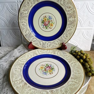 May include: Two vintage ceramic dinner plates with a white background, blue rim, and gold floral pattern. The plates feature a central floral design with pink, yellow, and blue flowers.
