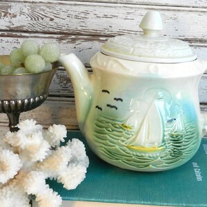 May include: A vintage ceramic teapot with a sailboat design, featuring a cream-coloured lid and handle, and a blue and green body. A silver bowl filled with green spheres and a white coral decoration are in the foreground.