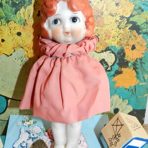 K&ouml;nnte beinhalten: Eine Vintage-Porzellanpuppe mit roten Locken, blauen Augen und einem rosa Kleid. Die Puppe hat ein detailliertes Gesicht und steht auf einer Oberfl&auml;che mit anderen Vintage-Artikeln, darunter ein Spielzeug-LKW und ein Block mit einer Drachenillustration.