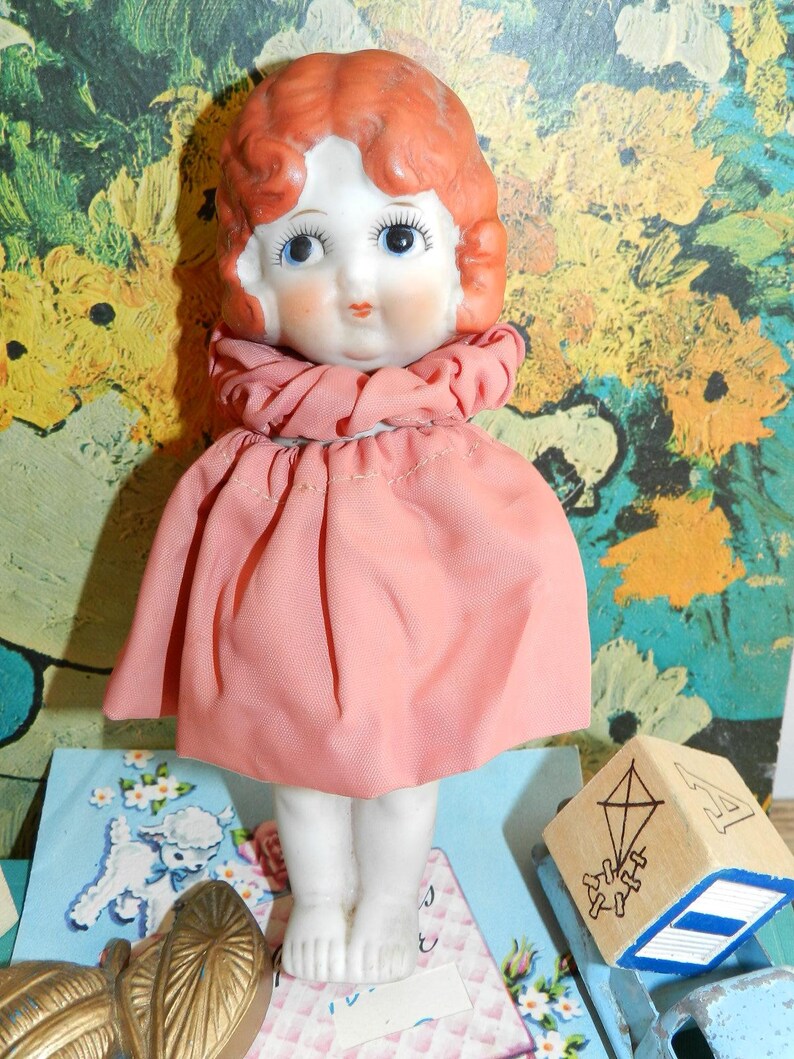 K&ouml;nnte beinhalten: Eine Vintage-Porzellanpuppe mit roten Locken, blauen Augen und einem rosa Kleid. Die Puppe hat ein detailliertes Gesicht und steht auf einer Oberfl&auml;che mit anderen Vintage-Artikeln, darunter ein Spielzeug-LKW und ein Block mit einer Drachenillustration.