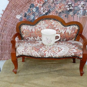 May include: A miniature wooden sofa with a floral patterned fabric upholstery. The sofa has a curved back and a cup sitting on the seat.